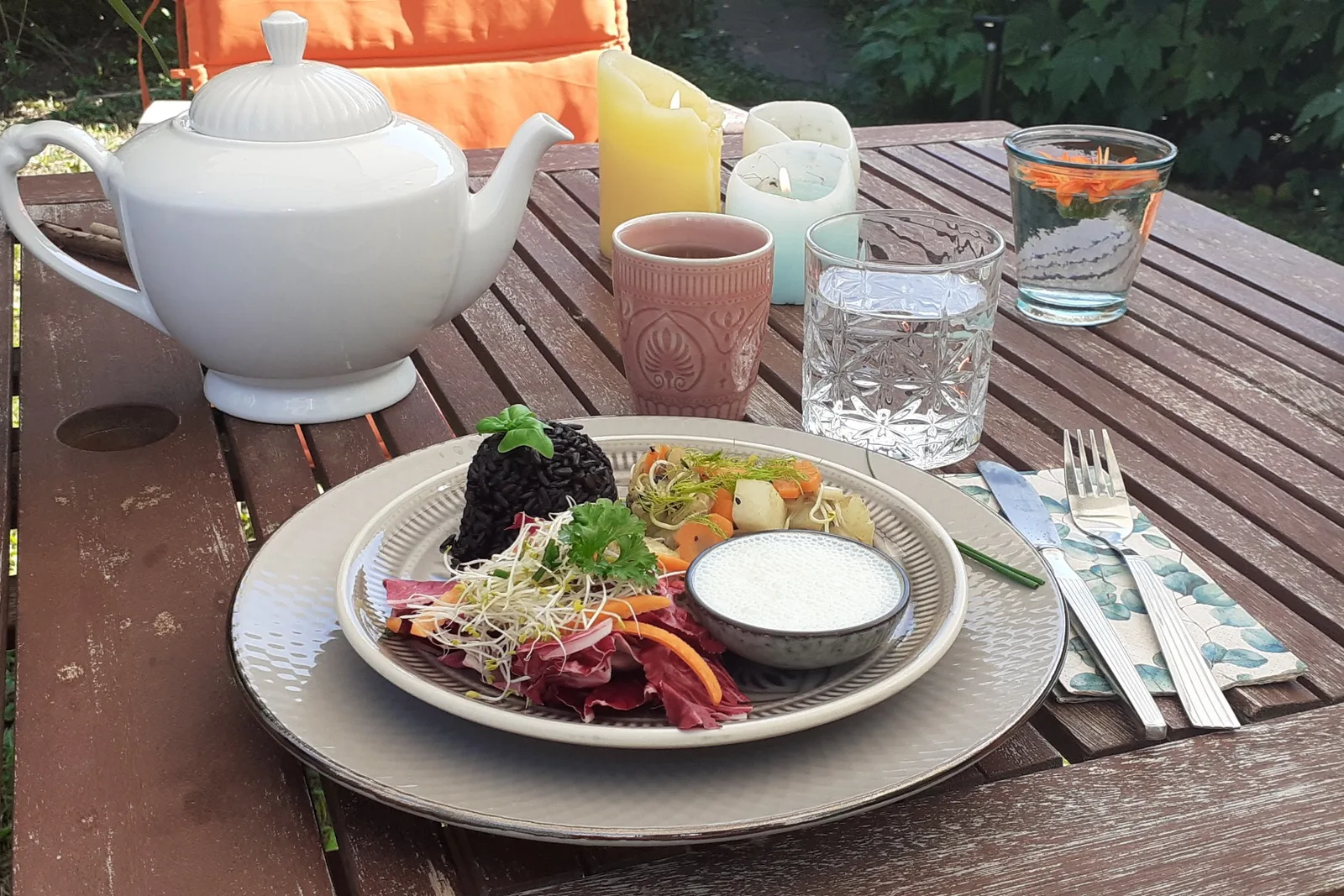 Ayurvedischer Teller mit schwarzem Reis, gedünstetem Fenchel-Kohlrabi-Möhren-Gemüse, buntem Sprossensalat und Gurken-Raita, seitlich fotografiert auf einem Holztisch im Garten